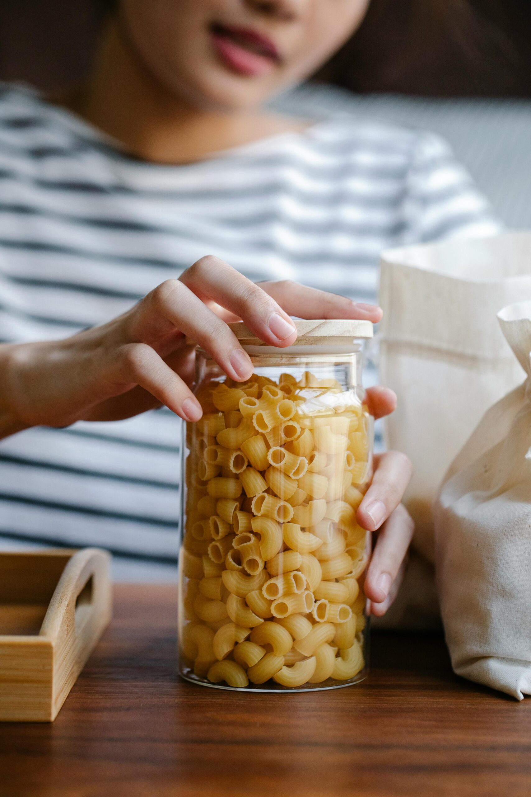 Crop anonymous female with transparent jar filled with pasta on wooden table in kitchen